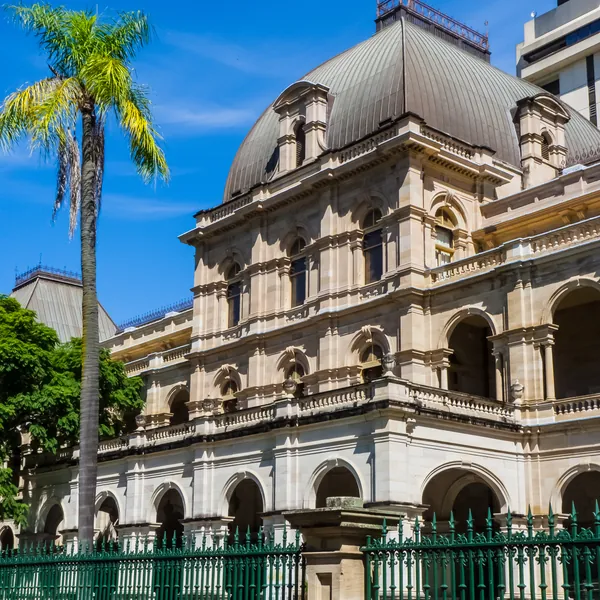 Queensland Parliament House on George Street: a sandstone corner pavilion with arched verandas and a tall copper dome behind a green iron fence, a palm beside it, under a blue sky